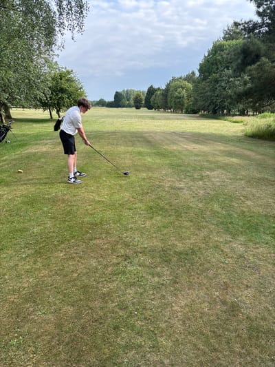 A young man in a white shirt and black shorts swings a golf club on a grassy field. The landscape includes green trees bordering the field under a cloudy sky. A golf bag is visible near the lower left corner. The field has a mixture of green and brownish grass. The man wears black and white shoes and has a golf glove on his left hand. The photograph is taken from a slightly low angle.
