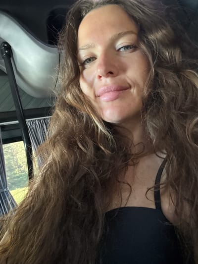 The image is a close-up selfie of a woman with long, wavy brown hair and fair skin. She is smiling and looking at the camera. She is wearing a black top. The background suggests she might be inside a vehicle or camper, with curtains visible to her left. A hint of green trees and foliage can be seen through the window, implying an outdoor setting, possibly near a rural area. There is no identifiable information that places her specifically in Turbaza Katun', Russia.