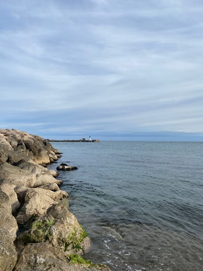 This image presents a coastal scene in Sète, France. The foreground and left side are dominated by a sturdy rock breakwater, composed of numerous irregularly shaped, light-colored boulders. Several rocks at the water's edge exhibit distinct patches of bright green algae. The sea, extending across the center and right, is calm with subtle ripples and a blue-grey coloration, revealing some submerged rocks near the shore. In the mid-distance, a pier or a further section of a breakwater extends horizontally into the water, ending with a small, prominent lighthouse. The lighthouse is white with a green top. The sky overhead is largely overcast, displaying layers of thin, horizontal stratiform clouds in shades of light blue and white. No individuals or specific human activities are visible.