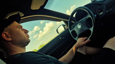This image depicts the interior of a car, likely a Volkswagen, seen from the perspective of the driver. The driver, a bald male wearing a black baseball cap and a dark t-shirt, is looking out the window. His left hand rests on the steering wheel. The car's dashboard is visible, including the speedometer and an odometer reading 32336. The exterior environment outside the car appears to be rural, with a sunny sky and clouds, and a field or landscape visible through the windows. The overall lighting suggests it is daytime. The location is specified as Mihailovca, Moldova.