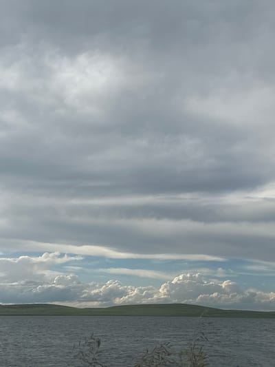 The image depicts a serene landscape with a body of water in the foreground, reflecting the overcast sky. The water is choppy, indicating a light breeze. In the middle ground, a gentle green hill slopes down to the water's edge. The sky is filled with layers of grey clouds, with some patches of lighter blue visible near the horizon. The overall atmosphere is calm and tranquil, suggesting a quiet day in the countryside. The scene could be located in or around Ozyorny, Russia, given its natural beauty and open spaces.