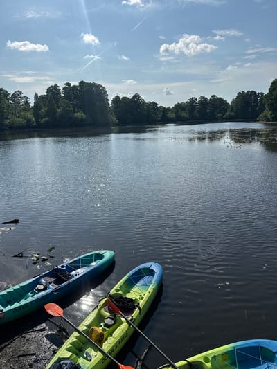 The image shows a body of water, possibly a lake or wide river, reflecting sunlight. Several kayaks are docked on the right side of the frame. The sky is partly cloudy with scattered clouds. Trees line the edge of the water on the left side of the image. There are no people visible. It is not possible to identify the precise location in Tampa with the data provided.