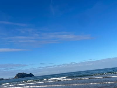 The image displays a coastal scene in Zarautz, Spain, facing the ocean. The upper two-thirds of the frame consist of a clear, vibrant blue sky interspersed with sparse, elongated white clouds horizontally oriented. The lower third features the dark blue waters of the Bay of Biscay, with several parallel lines of white-capped waves breaking and receding. In the left mid-ground, a dark, conical island, heavily vegetated, rises from the sea. No individuals, specific man-made structures, or direct interactions are visible. The environment is open-air and suggests daylight conditions.