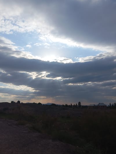 This image depicts a dramatic sky over a rural landscape. Dark, heavy clouds dominate the upper portion of the frame, with shafts of sunlight breaking through, creating a crepuscular ray effect. Patches of lighter blue sky are visible between the clouds. The sun itself is not directly visible but its presence is indicated by the intense light.
Below the sky, a dark, undulating terrain stretches across the middle ground. This appears to be a natural landscape with low-lying vegetation, possibly scrub or dry grass, and some scattered trees. In the distance, a few buildings are visible, suggesting a small settlement or scattered houses. These buildings have visible roofs and appear to be of residential construction. The overall impression is one of a somewhat remote, natural environment, possibly at dusk or dawn given the lighting conditions. The reference to the city of Kızıl-Bel, Kyrgyzstan, places this scene within a specific geographic context, suggesting a mountainous or hilly region with a dry climate. The foreground is a dark, rough-textured surface, likely a dirt road or unpaved ground, with sparse vegetation. There are no people or animals visible in the frame.