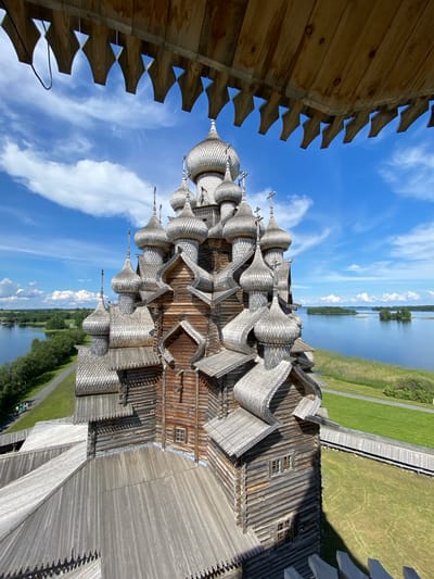 The image, taken from an elevated vantage point, shows the Kizhi Pogost, a historic architectural ensemble located on Kizhi Island in the Republic of Karelia, Russia, near the city of Boyarshchina. The primary subject is the Transfiguration Church, a UNESCO World Heritage Site, constructed entirely of wood. The church is characterized by its numerous onion domes, intricate wooden shingles, and tiered structure. Its weathered, natural wood exterior contrasts with the bright blue sky filled with scattered white clouds.
In the foreground, a wooden structure with decorative, notched eaves extends into the frame, suggesting the viewpoint is from a similar wooden building or observation deck. The church itself is situated on a grassy area adjacent to a body of water, which appears to be Lake Onega. The water's surface reflects the sky. Islands with green vegetation dot the distant water. A pathway, possibly a wooden walkway, runs along the edge of the grassy area and near the water. A few people are visible in the distance on a pathway to the left of the church, appearing as small figures. The overall scene depicts a serene, architectural landmark set within a natural landscape under a clear, sunny sky.