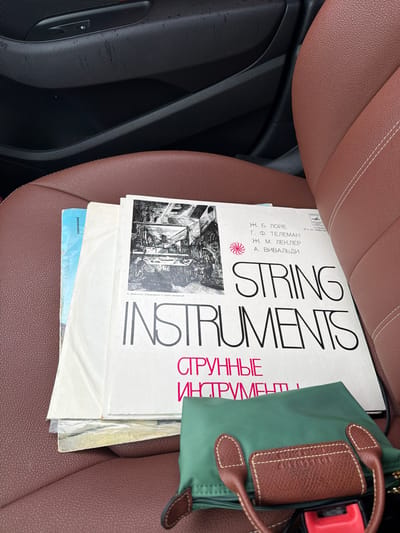 An aerial view of a car seat with two vinyl records. The record on top, with a white sleeve, displays the text "STRING INSTRUMENTS" in large black lettering, with "СТРУННЫЕ ИНСТРУМЕНТЫ" in smaller pink lettering below. The record sleeve also features a black and white illustration of a workshop with musical instruments. The second record, with a light blue sleeve, is partially visible beneath the first. The car seat is upholstered in brown leather with white stitching. A green fabric pouch with brown leather handles rests on the seat in front of the records. The background shows a black car door panel. The context of Darna, Russia is not discernable from the image.
