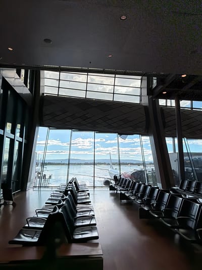 The image depicts an interior view of an airport terminal in Gardermoen, Norway. The scene is dominated by rows of empty, dark-colored waiting chairs arranged in a staggered pattern. Large windows offer a view of the tarmac, where an airplane is parked. The sky outside is partly cloudy, and mountains are visible in the distance. The terminal's architecture features a modern design with prominent structural elements and a high ceiling. The lighting suggests it is daytime. There are no people visible in the image.