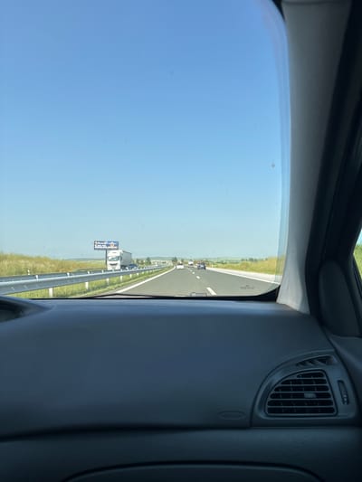 The image shows a view from the interior of a car, looking out through the front windshield onto a multi-lane highway. The car's dark grey dashboard, side pillar, and an air vent with "AIRBAG" text are visible on the right and top. The windshield, marked with "PILKINGTON," frames a bright, sunny daytime scene.
A clear blue sky dominates the upper left, while the highway stretches straight into the distance on the right, bordered by green, flat fields. Several vehicles, including cars and a truck, are driving on the highway. Guardrails line the edge of the road.
A prominent billboard by the roadside advertises "PRIMIRENT" with "РЕНТА-КАР" (RENT-A-CAR in Cyrillic) below it. Contact information includes phone numbers "0700 80 870" and "0886 070 870," and a website "primirent.com." The Cyrillic text and Bulgarian phone numbers are consistent with the provided location context of Malenovo, Bulgaria. The scene depicts travel on a highway under clear weather conditions.