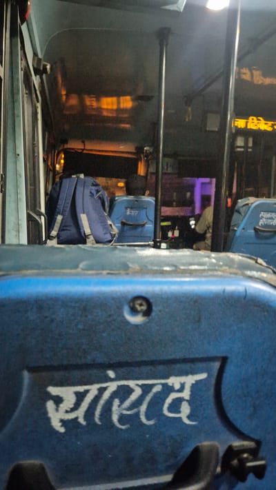 This image captures the interior of a bus at night, likely in Kullu, India, as suggested by the Hindi text. The focus is on the right side of the bus, revealing a blue panel with white Hindi lettering that appears to spell "आनंद" (Anand). To the left of this panel, a stack of luggage is visible. The top item is a blue rolling suitcase with a black handle, and below it is a blue backpack with grey straps. The luggage is positioned on what seems to be a luggage rack.

Further to the left, through the bus windows and internal dividers, the scene outside or further inside the bus is dimly lit. There are reflections on the window surfaces, hinting at interior lighting and possibly lights from outside. The overall atmosphere is dim, suggesting it is nighttime. The presence of luggage indicates passengers are likely traveling. The image is slightly angled, giving a dynamic perspective of the bus's interior.