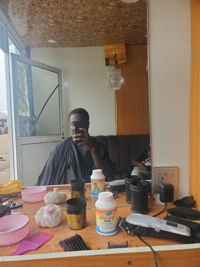In a barber shop, a young man is taking a selfie in a mirror while wearing a barber's cape. The mirror reflects a cluttered countertop with various hair cutting tools and products, including combs, clippers, powder containers, and puffs. In the background, a couch is visible, and a painting of a bird is hanging on the wall. The barber shop appears to be located in Ohit, Nigeria, as indicated by the signage on a container. The overall scene suggests a functional, albeit slightly disorganized, barbershop environment.