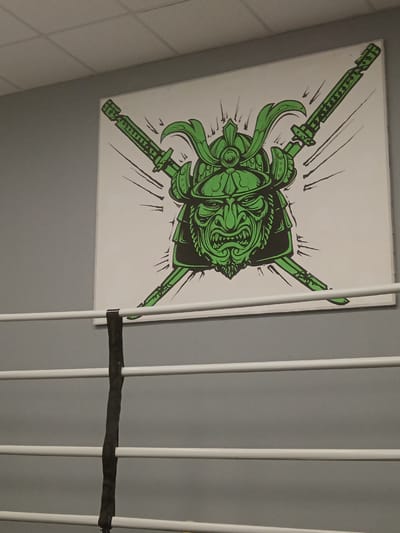 A framed painting of a green samurai mask with crossed swords hangs on a gray wall above three white horizontal bars, which appear to be part of a boxing ring. The painting is centrally located on the wall. The background of the painting is white with black lines radiating from the samurai mask. The samurai mask has a fierce expression with sharp teeth and intense eyes. The swords are also depicted in green with intricate details. A black strap hangs vertically between the white bars, partially obscuring the lower part of the painting. The ceiling above the wall is made of white acoustic tiles. The image is taken from a low angle, looking up at the painting and the boxing ring. There are no people visible in the image.