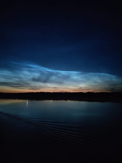 The image captures a serene twilight scene over a body of water, likely a lake or a wide river, under a dark blue sky. In the distance, a silhouetted treeline marks the horizon. Above the treeline, a band of luminous, ethereal clouds stretches across the sky, illuminated by the fading sunlight from below, giving them a pale blue and white glow. These are likely noctilucent clouds, a phenomenon that occurs at high latitudes during summer twilight.

In the water, a navigational buoy with a lit beacon stands as a solitary point of light. Emanating from the lower left of the frame are distinct, parallel ripples spreading across the dark surface of the water, suggesting the recent passage of a vessel or a disturbance from that direction. The overall impression is one of tranquility and natural beauty, characteristic of a remote, natural setting. The location specified is Zvoz, Russia, which is known for its natural landscapes and opportunities to witness phenomena like noctilucent clouds.
