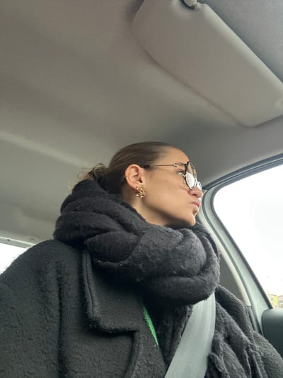 A single female subject is seated in the passenger seat of a vehicle in Saint-Cyprien, France. Her light brown hair is pulled back. She wears tortoiseshell-framed glasses, a small gold-colored stud earring in her upper left ear cartilage, and a larger, intricate gold-colored cluster earring in her left earlobe. She is dressed in a thick, textured black coat over a bright green garment, partially obscured by a voluminous, fringed black scarf wrapped around her neck and shoulders. A light grey seatbelt is visible across her chest and right shoulder. Her head is turned towards the right, looking out the vehicle's window, with her lips puckered. The vehicle's interior features a light grey headliner and a matching sun visor mounted above the passenger seat. The exterior visible through the window is a bright, overcast sky.