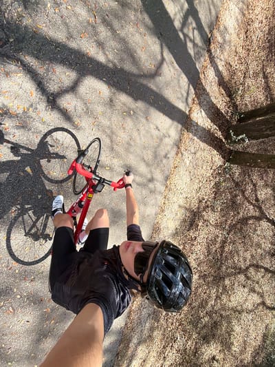 An overhead photograph depicts an individual on a red road bicycle stopped on a paved path in Gainesville, United States. The person, wearing a black cycling helmet, dark sunglasses, a black textured cycling jersey, black cycling shorts, white cycling shoes, and a watch on their left wrist, holds the camera overhead while gripping the bicycle's black handlebars, which are wrapped in red tape. A small electronic device is mounted on the handlebars, and a white water bottle is secured in the bicycle's frame. The asphalt path is partially covered with scattered brown and orange fallen leaves. To the right, an unpaved area is heavily strewn with similar leaves, bordered by visible tree trunks. Strong, direct sunlight casts long, distinct shadows from trees and the bicycle across the scene, including the rider's shadow on the pavement.