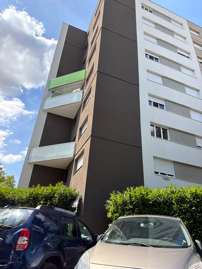 The image displays a modern apartment building with a dark grey facade and light beige sections featuring vertical white blinds and windows. The building has protruding balconies with frosted glass panels, one of which is a distinctive green color. In the foreground, partially visible cars are parked, one a silver sedan and another a dark blue SUV with "DUSTER" written on its rear. Lush green bushes flank the building and cars, suggesting an outdoor setting. The sky is a clear blue with scattered white clouds, indicating fair weather. The upward angle of the shot and the presence of cars and vegetation suggest an urban or suburban environment. The lighting implies it is daytime. No people are visible. The image is taken from a slightly tilted perspective.
