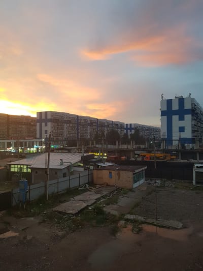 The image depicts a cityscape at dusk, with a notable presence of residential buildings. In the foreground, a muddy, unpaved area with scattered debris and a few small structures is visible. Beyond this, several elongated, multi-story apartment buildings line the horizon. The architecture of these buildings appears to be consistent, with repeating balcony structures and a similar color scheme of light facades and blue accents. One prominent building features a large blue cross-like symbol on its facade. Construction activity is evident in the mid-ground, with scaffolding and incomplete structures suggesting ongoing development or renovation. The sky is a canvas of twilight colors, with soft hues of orange, pink, and blue, indicative of sunset or sunrise. The overall impression is one of a developing urban environment in Бұлақты, Kazakhstan, characterized by mass housing and ongoing construction.