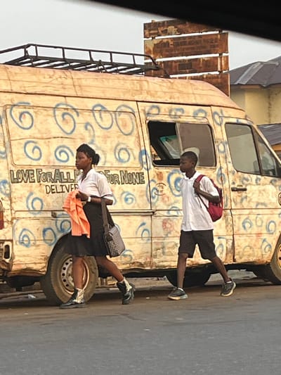 The image captures a street scene featuring a distinctive, old-model panel van and three individuals during what appears to be daytime. The van is painted a faded cream or off-white color and is extensively decorated with numerous blue, swirling, circular patterns across its side. The paint shows signs of wear and chipping, indicating the vehicle's age and frequent use. A black metal ladder is affixed to the left side of the van, leading up to a roof rack which holds several wooden planks or pallets.
Two individuals are standing on the asphalt road next to the van's passenger side. Closer to the front of the van is a young male wearing a white short-sleeved shirt, dark shorts, and sneakers. He is carrying a red backpack and has his back partially turned towards the camera, looking towards the van. Further back, near the rear wheel of the van, stands a young female. She has dark hair styled in braids or dreadlocks, pulled back. She is dressed in a white short-sleeved top, dark shorts or a skirt, and patterned sneakers. She is wearing a gray patterned shoulder bag and holds an orange cloth or garment in her hands, facing mostly towards the van.
Inside the van, a third individual's head is visible through the open driver's side window, looking forward or slightly downward.
Prominently displayed text painted on the side of the van reads: "LOVE FOR ALL HATRED FOR NONE". Below this, another word, partially obscured, appears to be "DELIVERY" or "DELIVERING". The ground is a dark, asphalt-like surface typical of a street or road. The overall scene suggests a moment of casual waiting or a pause in activity, possibly related to a transport or delivery service.