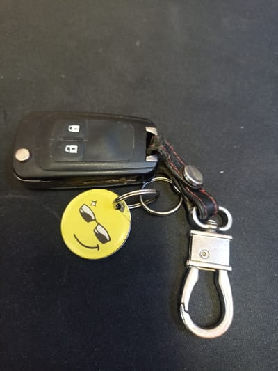 This media file shows a close-up, eye-level view of a car key fob with a keychain attached. The key fob is black and appears to be made of plastic, with two buttons visible that have symbols of a closed and an open padlock. There is a silver circular button on the left side of the fob. Attached to the fob by a leather strap with red stitching and a silver stud is a round yellow charm. The charm features a cartoonish smiley face wearing sunglasses, with small sparkle marks above the sunglasses. A silver keyring connects the charm to the leather strap. Below the charm and strap, a silver carabiner-style clip is attached, which would be used to fasten the keys to a belt loop or bag. The objects are placed on a dark, textured surface, likely a table or fabric, which is dimly lit. There is no indication of the time of day, weather, or specific location beyond the context provided. The overall mood is casual and lighthearted due to the smiley face charm. There is no visible text in the image.