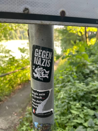 The image presents a close-up, eye-level view of a grey metal utility pole situated outdoors, characteristic of a park or natural pathway in Schongau, Germany. At the top of the pole, a portion of a white, reflective rectangular sign with a horizontal black stripe is attached by two visible flathead screws; the number "66" is partially visible on the sign's upper right corner.

Affixed to the pole are multiple stickers. The uppermost prominent sticker is rectangular with a black background and white text that reads "GEGEN NAZIS" (Against Nazis). Below the text, a white graphic depicts a clenched fist crushing a swastika symbol. This sticker shows signs of wear, including minor tears and surface abrasions. Directly beneath it, a partially peeled and damaged black sticker displays white text, with discernible phrases such as "Single sein ist Dreck..." (Being single is dirt/crap...) and "Lass dein Freund wisch..." (Let your friend swipe...). A Facebook logo and fragments of German text, including "Freundinnen verkuppeln" (matchmaking friends), are also visible. The lowest visible sticker fragment shows a partial image of a wooden structure amidst green foliage.

The background scenery suggests a natural environment. To the left of the pole, a body of water, likely a lake or pond, is visible, reflecting bright sunlight. A narrow dirt path or trail runs along the water's edge, bordered by green trees and bushes. To the right, dense green foliage, primarily shrubs and trees, occupies the background, with sunlight filtering through the leaves, creating a blurred, luminous effect. No individuals or overt actions are depicted.