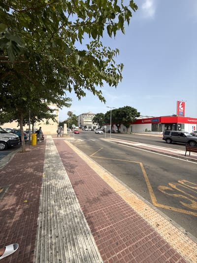 The image shows a street scene in Inca, Spain, with a focus on the sidewalk and the road. On the sidewalk, there are several people sitting on a bench and a yellow trash can. The sidewalk is paved with reddish-brown tiles and has a tactile paving strip. Trees with green foliage overhang the sidewalk. Across the street, there is a red building with a sign that reads "EROSKI center Supermercado." Several cars are visible on the road, including a gray SUV and a white van. The sky is blue with some clouds. The environment appears to be an urban setting on a sunny day.