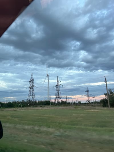 This image depicts a landscape with power lines and a wind turbine under a cloudy sky. The scene appears to be a rural or semi-rural area, with a large grassy field on the right side of the frame and trees lining the horizon. Several tall power transmission towers, characteristic of those found in Zimovniki, Russia, are visible, along with the distinctive white blades of a wind turbine.
The time of day is likely dusk or dawn, suggested by the soft, diffused light and the pastel hues in the sky between the clouds. The clouds themselves are a mix of dark grays and lighter blues, indicating potentially overcast or changeable weather conditions. The overall mood is somewhat somber due to the heavy clouds, but the presence of the wind turbine also hints at a modern, perhaps even hopeful, energy infrastructure.
No people are visible in the image. The primary subjects are the power transmission infrastructure and the natural elements of the sky and field. The image seems to be taken from a moving vehicle, as suggested by the slight blur in the grassy field and the dark, out-of-focus object in the bottom left corner which could be part of the vehicle's interior. There is no visible text in the image.