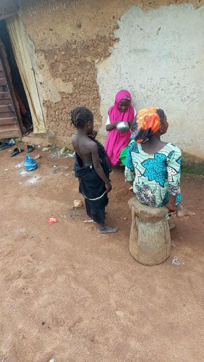Three young girls are gathered outside a rustic dwelling in Kagarko South, Nigeria. The building appears to be constructed from mud and stone, with a rough, textured wall. A tattered curtain hangs over an open doorway to the left. The ground is a dusty, reddish-brown earth, with scattered pebbles and debris.

In the foreground, a girl with dark, braided hair is facing away from the camera, her back bare. She wears a dark, flowing garment that drapes around her legs, reaching her ankles. She has a small hoop earring in her visible ear.

To her right, a girl in a bright pink hijab looks down and holds a silver bowl. She is dressed in a white top beneath the hijab and the pink outer garment.

Further to the right, a third girl sits on a roughly carved wooden stool. She wears a colorful long-sleeved dress with a vibrant floral pattern in shades of blue, green, and black. Her head is covered with an orange and black patterned headscarf. Her right foot is visible, resting on the stool.

Scattered on the ground are several objects: a pair of black flip-flops, a blue plastic container with a handle, and a small, red rectangular object that appears to be a packet of sweets. A piece of white string is also visible on the ground. The overall scene depicts a simple, outdoor domestic setting.