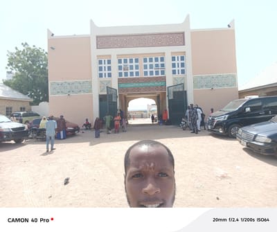 The image is a selfie taken in front of a building with architectural features including geometric patterns, windows, and an arched gate, identified as being in Wase, Nigeria. The building is peach-colored, with geometric patterns in brown and blue above and below the windows. Arabic script adorns the building above the gate. People are standing and walking around. There are vehicles parked to the left and right of the building. The ground is sandy and dusty. A pigeon stands on the ground in front of the photographer.