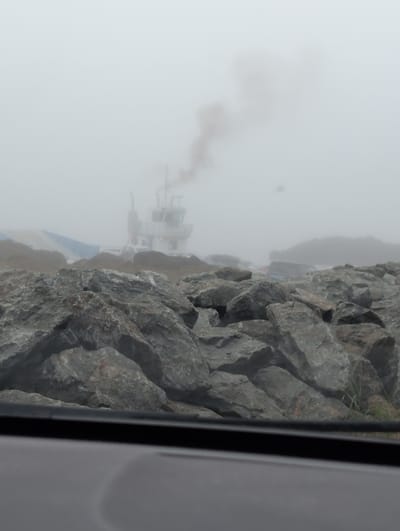 A tugboat with smoke billowing from its stack is partially visible through thick fog, in the waters near West Isles Parish, Canada. The foreground is dominated by a jumble of large, grey rocks. In the background, a portion of the tugboat's white superstructure and a blue and white striped hull are discernible through the mist. The sky is overcast and uniformly grey.