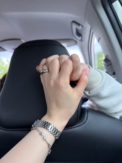 Inside a vehicle, two individuals are holding hands. One hand is adorned with a silver ring on the ring finger, a metal watch, and a star bracelet. The other hand is wearing a white, long-sleeved sweater. The location is in Tserovani, Georgia, as evidenced by the setting. The scene appears to be captured during the day, with natural light entering the vehicle.