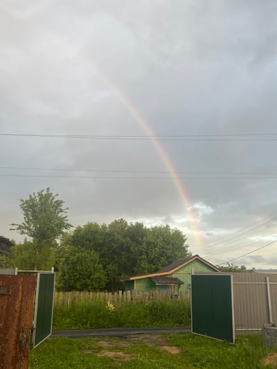 The image depicts an outdoor scene in Klobushnevo, Russia, under a cloudy sky with a visible rainbow arcing across it. In the foreground, a weathered, partially open gate with peeling red paint stands on the left, revealing a glimpse of green grass and a dirt path. To the right of the path, a tall, dark green gate is open, leading towards a fence made of vertical wooden posts topped with horizontal planks. Beyond the fence, a small, green wooden house with a brown roof and trim is visible. Dense green trees form a backdrop behind the house and fence. Power lines are strung horizontally across the sky, with one set lower than the other. The overall impression is of a rural or suburban setting on a day with recent or ongoing rain. There are no people or animals visible in the image.