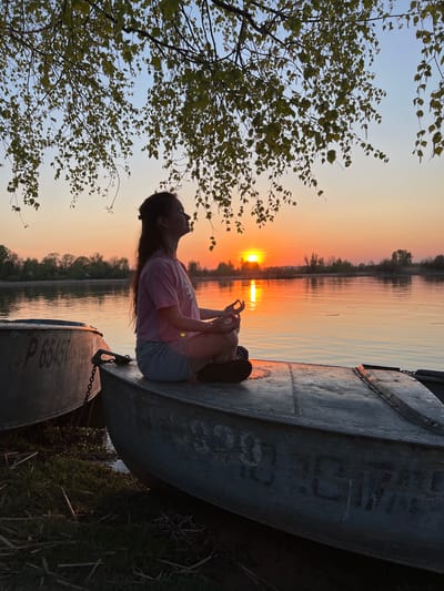 A long-haired woman is sitting in a metal boat at sunset. The location is most likely near Borovoye Matyushino, Russia. She's wearing a pink shirt and denim shorts. The sun is reflected on the water behind her. In the bottom right corner and bottom left corner is another boat. A tree is situated on the left-hand side.