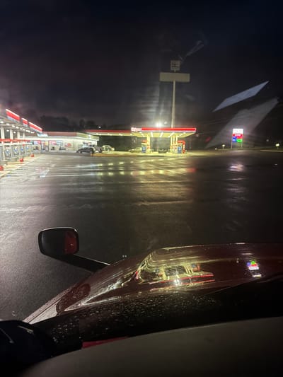The image depicts a view from inside a vehicle at night, looking out towards a gas station. The ground is wet and reflects the lights of the gas station and nearby buildings. A red truck's hood is visible in the foreground, with raindrops on its surface. The gas station has red and white awnings covering the pumps, which are illuminated. A sign with "Exxon" is visible. There are several cars parked at the gas station, and a few indistinct figures are present. The sky is dark with some clouds. The scene appears to be a typical gas station illuminated at night, likely after or during rainfall, in the city of Wildwood, United States.