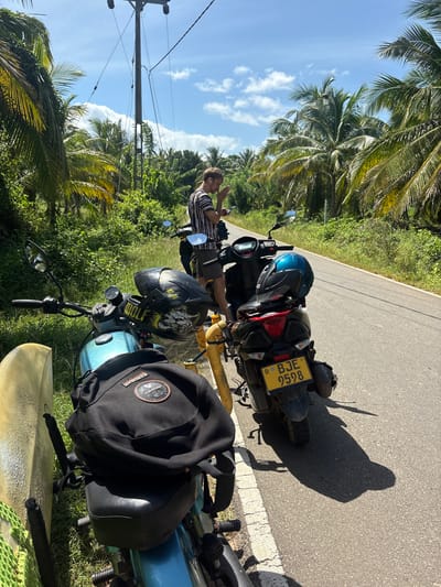 A man stands by a scooter on the side of a paved road in an area with lush green vegetation and palm trees. The man is wearing a striped t-shirt and dark shorts, and is looking down at his phone. Two scooters are parked side-by-side, both with helmets placed on the seats. A surfboard is leaning against the left scooter. The scooter on the right has a yellow license plate with the registration number BJE 9598. The sky is blue with scattered clouds. The overall scene suggests a stop during a journey in a tropical location in Sri Lanka.