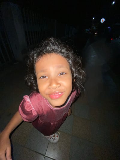 A young girl with curly dark hair is looking up at the camera in what appears to be a residential area at night. She is wearing a dark pink velvet dress with ruffled sleeves and white shoes. Her mouth is slightly open, revealing her teeth. The background is dark, with some faint lights and the suggestion of a fence or wall on the left side. The ground is paved with rectangular tiles. The image is taken from a low angle, looking up at the girl. While the specific location in Tanggul, Indonesia, is not identifiable from the image itself, the context of a residential area at night with paved ground is consistent with many urban and suburban environments.