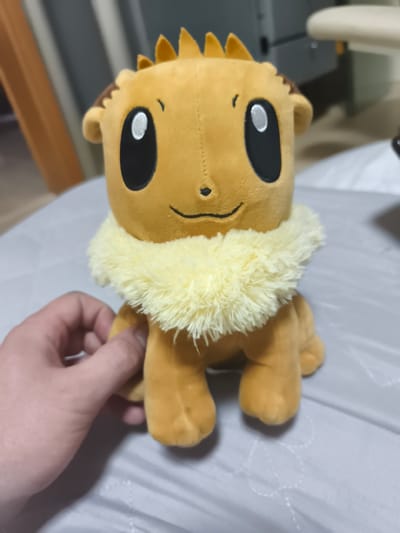 A plush toy, resembling the character Eevee from the Pokémon franchise, sits on a grey, textured surface. The toy is primarily light brown with a fluffy, pale yellow mane around its neck. Its large, black eyes have white pupils, and a small, black nose is centered above a stitched smile. A few short, spiky brown tufts protrude from the top of its head. A person's hand, with visible fingers and nails, is gently holding the toy from the left side, near its body. The background is blurred, showing indistinct shapes and colors, suggesting an indoor setting. The lighting appears to be artificial, casting soft shadows. The overall mood is calm and perhaps playful, given the presence of the toy. There is no visible text in the image.