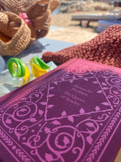 In Panormos, Greece, a close-up shot focuses on a book titled "The Queen of Spades" by Alexander Pushkin, resting on a table outdoors. The book is deep purple with intricate pink floral patterns and decorative borders. Nearby, two brightly colored plastic hair clips, one green and one yellow, are visible. In the background, out of focus, are woven straw bags and what appears to be a beach setting with lounge chairs and sand. The scene suggests a relaxed, leisurely day at the seaside.