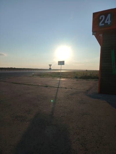 The image depicts an outdoor scene at sunset. In the foreground, there is a paved area, possibly a parking lot or roadside. To the right, a portion of a building with dark wood siding is visible, featuring a large white number "24" against an orange background. The building appears to be a commercial establishment, potentially a roadside service or shop.
In the mid-ground, a road stretches horizontally across the frame, with a guardrail along its edge. Several road signs are present: a triangular warning sign and a circular blue sign with a white arrow indicating a turn. Further in the distance, the sun is setting, casting a bright glare that partially obscures the horizon. A body of water, likely a river or lake, is visible beyond the road, with vegetation along its banks. The sky is a clear blue, transitioning to lighter hues near the horizon. The overall impression is that of a quiet, rural or semi-urban setting at the end of the day. The location is identified as being in the city of Arshaly, Kazakhstan.