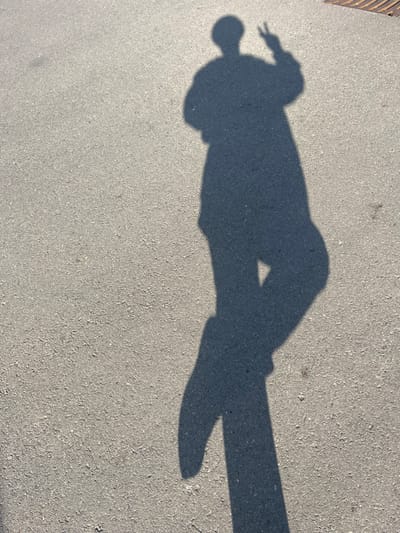The image displays the dark shadow of an individual cast upon a light grey, granular paved surface, likely asphalt or concrete, located in Roodt-sur-Syre, Luxembourg. The shadow, originating from an unseen person positioned to the right and slightly behind the camera's perspective, occupies the right two-thirds of the frame. The figure's shadow shows the person facing left, with their head, torso, and legs visible. The person's left arm (appearing on the right side of the shadow) is raised, with the hand forming a "V" gesture, commonly recognized as a peace sign. The right arm's shadow is visible hanging down the body. The legs are depicted as slightly separated, with the left leg (right side of the shadow) appearing straight and the right leg (left side of the shadow) bent at the knee, suggesting a relaxed posture or slight movement. The ground surface exhibits minor irregularities and faint marks. A small section of a dark metal grate or drainage cover is visible in the upper right corner of the frame. The distinct, sharp outline of the shadow indicates bright, direct sunlight.