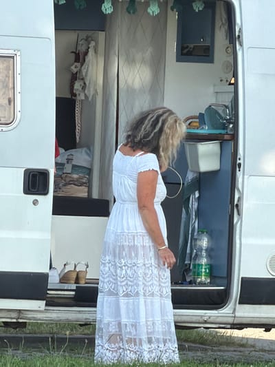 A woman in a white lace dress stands in the doorway of a white camper van. The van's interior is visible, featuring a blue cabinet, a sink, and various items. Outside the van, on the ground, are a pair of tan wedge sandals and a white shoe. The scene appears to be set in a grassy area, possibly near a parking lot or campsite, and is located in Ribnitz-Damgarten, Germany.