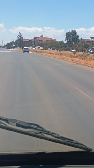 A wide, empty road stretches into the distance under a sunny sky. Buildings and trees line the roadside, with a few cars and pedestrians visible in the distance. The road surface is grey, and the verge is dry and reddish. The lower portion of the image shows the windshield and wipers of a vehicle from which the picture is taken.