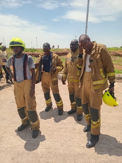 The image depicts four men wearing fire-fighting uniforms. One of the men, who is holding a yellow helmet in his left hand, is wiping his eyes with his right hand. The men are standing on a dirt road, with a light-colored paved area on their left. The background shows a clear blue sky with white clouds, and a field of grass. They are standing at approximately 10.71 Latitude and 7.514 Longitude in Katabu, Nigeria.