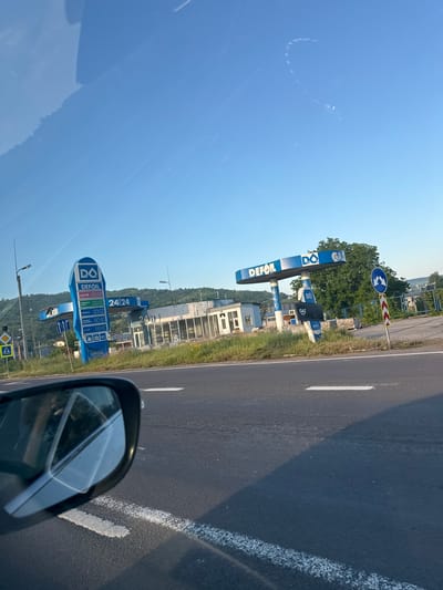 The image depicts a roadside scene at a gas station in an unknown location in Moldova, likely during daylight hours due to the bright sky. The primary focus is on a "D(q) Defoil" branded gas station with multiple pumps visible. One prominent blue sign displays fuel prices, including "SUPER 95" at 8.88 and "GAZ" at 2.88, indicating it's a 24-hour establishment. The gas station structure appears somewhat weathered and partially abandoned, with an adjacent building exhibiting signs of disrepair. A blue and white road sign with a mountain graphic and a red and white chevron patterned post are positioned to the right of the gas station. A yellow pedestrian crossing sign is visible on the left. The perspective is from inside a vehicle, as suggested by the reflection in the side mirror. The road in the foreground is asphalt with white lane markings. Greenery, including grass and trees, surrounds the gas station, with a hill or mountains in the background under a clear blue sky.