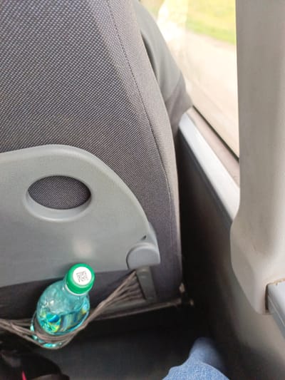 The image captures a close-up view of a blue plastic water bottle with a green cap, resting in a fabric netting pocket attached to the back of a gray bus seat. The bottle is partially filled with water, which reflects colorful light. On the cap, a small QR code is visible. The seat appears to be made of textured fabric. To the right of the seat, a portion of a window is visible, showing a blurred green and yellow outdoor scene, suggesting the bus is in motion. In the lower right corner, a person's blue jeans-clad leg is visible, indicating a passenger is seated. The overall scene suggests a moment of travel. The location is specified as Selo Lugovoye, Russia.