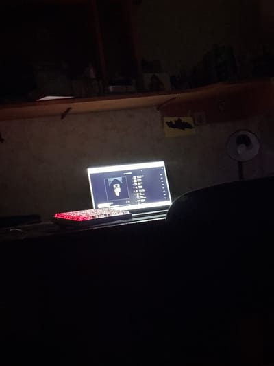 This image, contextualized in Lyman, Ukraine, captures a dimly lit indoor scene dominated by a glowing electronic screen. The rectangular screen displays a digital interface featuring a row of small white squares at the top, a bar graph or audio equalizer below, and a clear graphic of a clenched fist gesture. To the right of the screen, an electronic device with a series of illuminated red components, possibly LEDs or circuitry, is visible.

On the wall to the left, a dark silhouette of a bat is affixed, possibly a decorative element. Above it, a wall-mounted light fixture is present but unlit, further emphasizing the low light conditions. The surrounding environment is mostly shrouded in darkness, with indistinct shapes of furniture or shelving barely discernible on the far left. The right side of the image fades into complete blackness. The scene appears to be set at night or in a room with minimal ambient lighting. No human subjects or direct activity are visible; the focus is on the illuminated screen and electronic components, suggesting a setup for monitoring or display. No discernible text is legible on the screen.
