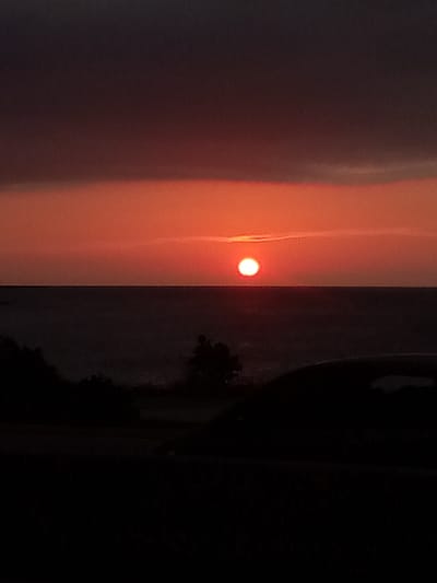 A sunset is captured over the ocean with dark clouds in the upper portion of the sky and a bright orange glow emanating from the horizon. The sun, a small, brilliant white circle, is visible just above the water. In the foreground, silhouetted against the colorful sky, is a dark shape of a car, partially obscuring a dark line of trees and vegetation. The scene is set at dusk, creating a dramatic and moody atmosphere. The location is identified as La Guaira, Venezuela.