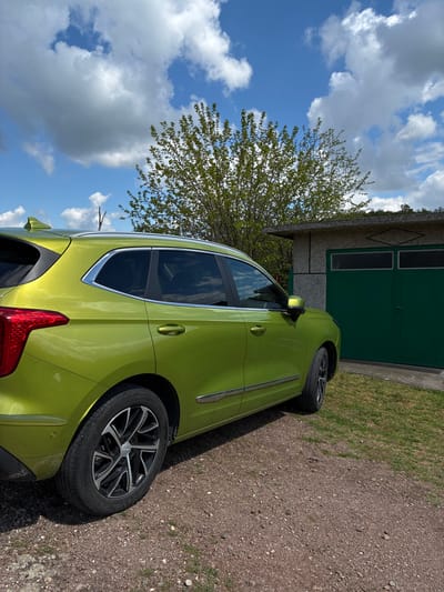 The image shows a light green car parked on a gravel surface near a small, single-story building with green doors. The building appears to be a garage or storage shed. A tree with budding green leaves stands near the car. The sky is blue with scattered white clouds. The location is implied to be Bokilovtsi, Bulgaria. There are no people visible in the image. The primary subject is the car, its color, and proximity to the building. The environment suggests a rural or suburban setting.