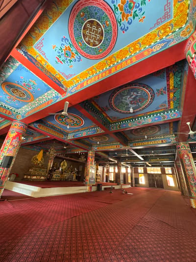 The image depicts the interior of a Buddhist temple in Việt Trì, Vietnam. The ceiling is elaborately decorated with vibrant colors and intricate patterns, featuring motifs such as lotus flowers, mythical creatures, and Tibetan knots. Red beams and pillars support the ceiling, which also has circular painted medallions and decorative trim. Several ceiling fans are visible, indicating the need for climate control.
The floor is covered with a red carpet featuring a geometric pattern of interlocking diamonds. In the background, a raised altar is adorned with statues of Buddha and other religious figures, draped with yellow fabric. Numerous columns, painted with colorful designs similar to those on the ceiling, support the structure. Large windows are present in the background, allowing sunlight to stream into the temple, creating bright illumination in parts of the room. The overall impression is one of a richly decorated and spiritual space.