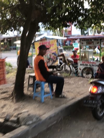 A person is seated on a blue plastic chair in front of a food stall. The person is wearing an orange and black shirt and has dark hair. Behind the person, the food stall has a yellow banner with some text and graphics. In the background, there are more stalls, some motorbikes, and trees. The scene appears to be outdoors, possibly on a street or a market. The image is blurry, suggesting motion or a low-quality capture.

The location is Sidoarjo, East Java. The image seems to be taken during the daytime, given the natural lighting. The overall atmosphere appears casual and busy, typical of a street-side food vendor setting in Indonesia. No specific emotion is discernible due to the blurriness.

Visible text: The yellow banner has some text in what appears to be Indonesian, but it is too blurry to read clearly. There is also text on other signs in the background that are equally unreadable. A motorcycle license plate is partially visible on a motorbike in the background, but the characters are illegible.
