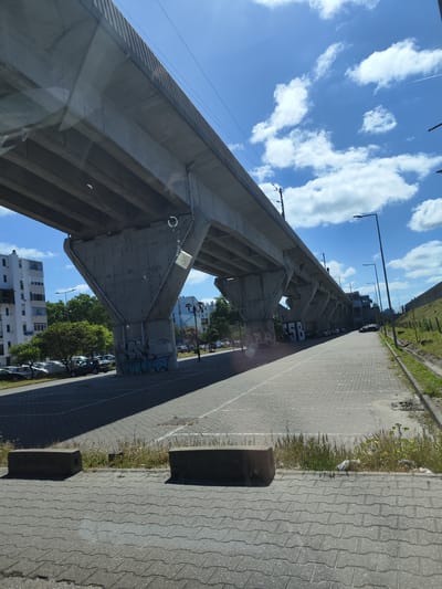 An outdoor, eye-level shot captures a bright urban scene in Seixal, Portugal, dominated by a massive elevated concrete structure, likely a viaduct for a railway or highway, extending diagonally from left to right. The structure is supported by heavy concrete pillars, some of which feature graffiti. One prominent pillar clearly displays "5B" in large white characters, while another further left has a more abstract, blue and black graffiti design.
Below the elevated structure, a paved area, resembling cobblestones, stretches into the distance, with strong shadows cast by the pillars and the overhead structure, indicating a sunny day. To the left, several cars are parked in an open lot under trees, next to white apartment buildings with balconies. To the right, the paved area borders a strip of wild grass and a small dirt embankment, with streetlights lining the path.
The sky is a vibrant blue with scattered white cumulus clouds. High in the sky, a faint contrail from an airplane is visible. The sun is high, creating sharp contrasts between sunlit areas and deep shadows. The image appears to be taken from inside a vehicle, evidenced by subtle reflections on the glass in the foreground, including a circular shape and faint streaks. Two concrete blocks rest on the paved ground in the bottom foreground, marking the edge of a grassy strip.