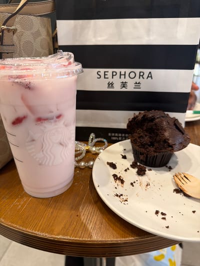 An overhead shot captures a collection of items on a wooden table, suggesting a casual break or outing. The scene features a partially eaten chocolate muffin, a pink beverage in a Starbucks cup, a Sephora shopping bag, and a beige purse.
The dark chocolate muffin, still in its paper liner, rests on a white ceramic plate. A portion of the muffin has been eaten, leaving crumbs scattered across the plate, alongside a small, light-colored wooden fork. Adjacent to this, a clear plastic Starbucks cup contains a light pink, opaque drink with ice and visible red fruit pieces, likely strawberries. The iconic Starbucks siren logo is clearly visible on the cup's surface.
Behind these items stands a black and white striped Sephora shopping bag. The brand name "SEPHORA" is printed vertically in capital letters on a white stripe, immediately followed by the Chinese characters "丝芙兰" (Sīfúlán), the brand's name in Chinese. To the left, a beige or light brown purse with a patterned texture and a gold-toned buckle is partially visible. A piece of shimmering jewelry, possibly a pearl or rhinestone necklace, is also visible on the table.
The setting appears to be indoors, with a wooden tabletop featuring a distinct grain. The presence of the Sephora bag with Chinese text strongly indicates the location is in China, aligning with the provided context of Yuhuatai District. The image portrays a moment of relaxation, possibly after a shopping trip.